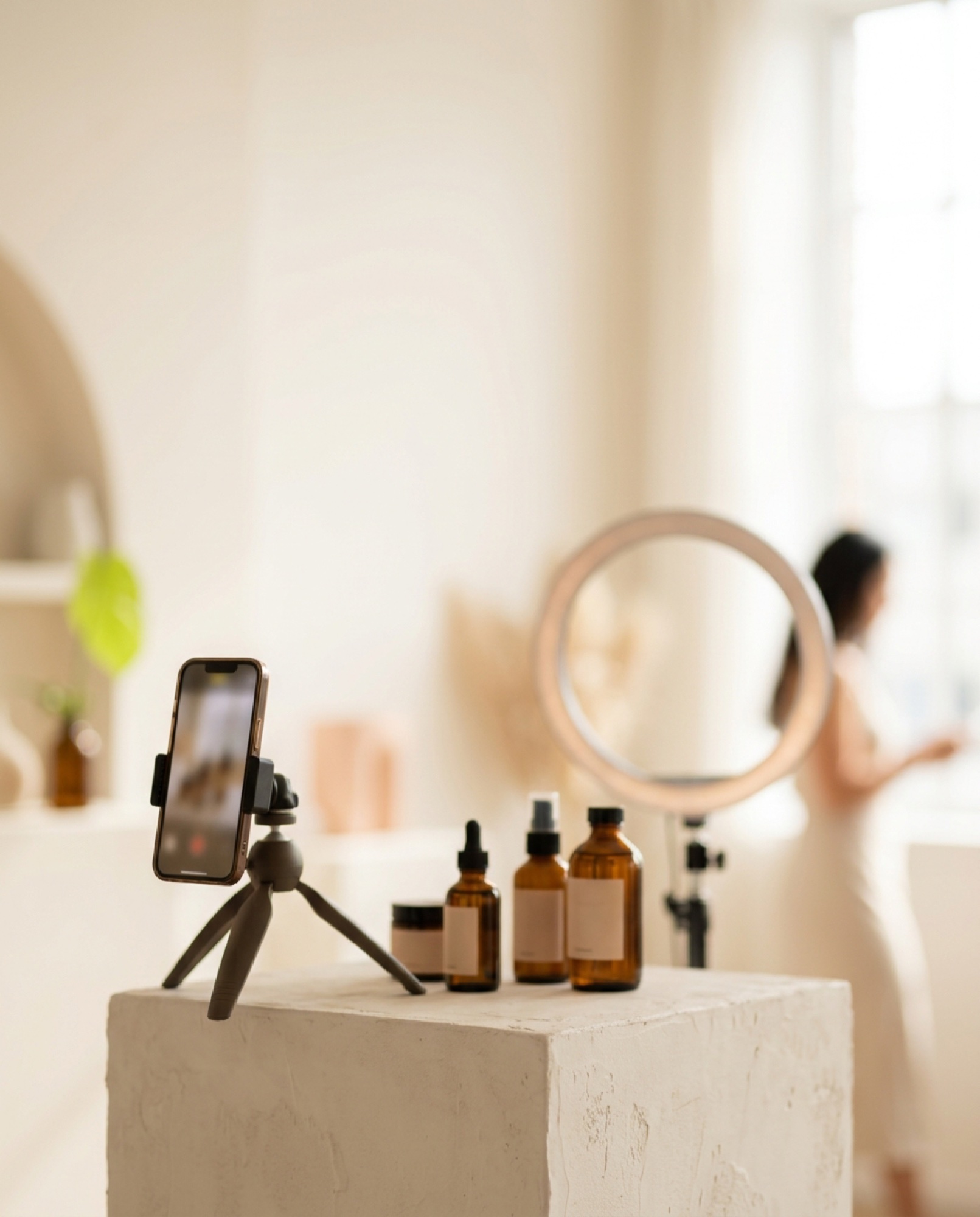 A phone on a tripod filming skincare bottles in a sunlit studio, a creator by a ring light behind
